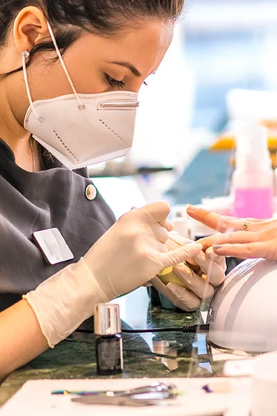 A person wearing a mask and gloves is focusing on performing a manicure on another person's hand. Various nail care tools and products are visible on the table, including nail polish bottles.