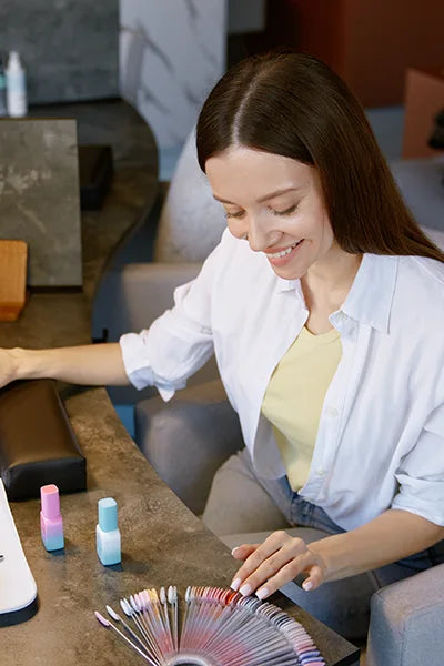 Woman with long hair selects a nail polish color from a palette at a salon. She is smiling and sitting at a table with nail polish bottles nearby. She's wearing a white shirt over a yellow top.