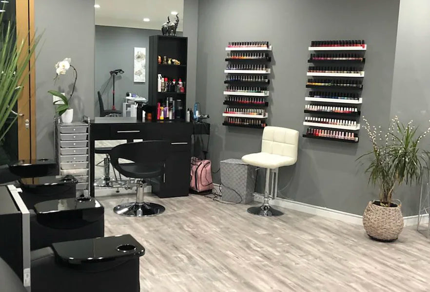 A modern nail salon with gray walls and wooden flooring. Shelves with organized nail polish bottles line the wall. Two black chairs face a counter with beauty products. A white stool and potted plants add to the decor.