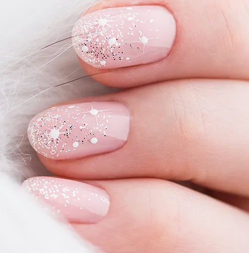 Close-up of a hand with manicured nails, painted in pale pink with silver glitter and star patterns. The person is wearing a fluffy white garment, partially visible in the background.