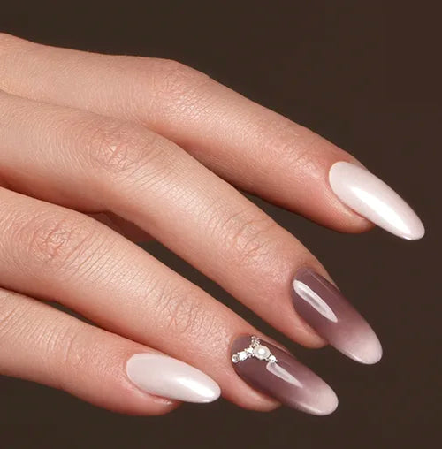 A hand with long, almond-shaped nails. The nails are painted in a gradient from light pink to dark purple, and one nail has small, sparkling rhinestones as decoration. The background is dark brown.