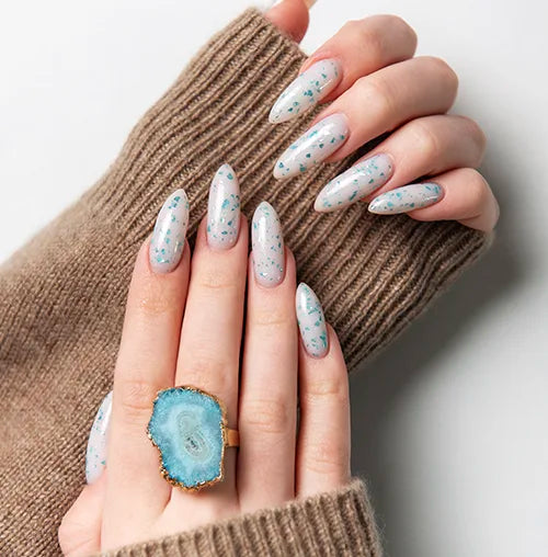 Hands with long, oval-shaped nails painted in a light blue shade with small blue speckles. The person is wearing a beige sweater and a ring with a large, round blue stone.