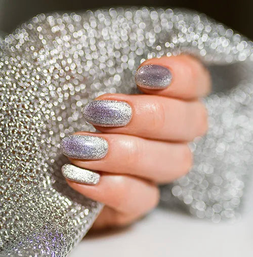 A hand with silver glittery nail polish gently grasps a sparkly, textured silver fabric. The lighting highlights the shine and shimmer of both the nails and the fabric.