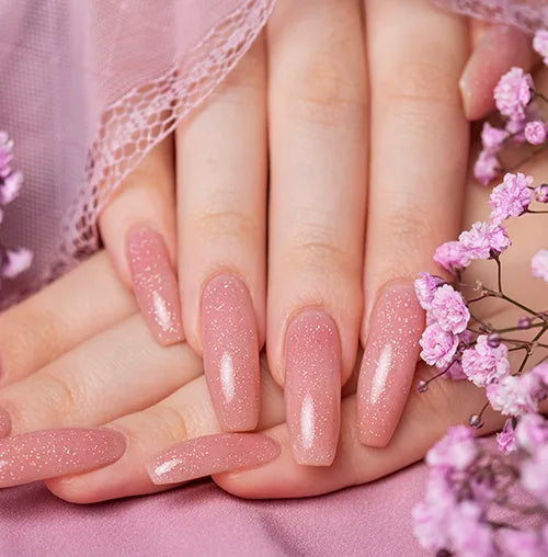 Close-up of hands with long, glittery pink nails surrounded by pink flowers. A delicate pink lace fabric partially covers the hands, enhancing the soft, feminine aesthetic of the scene.