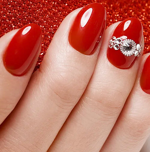 A hand with neatly manicured nails painted in glossy red polish. One nail is adorned with elegant silver rhinestone decorations, and the background features a sparkly red surface.