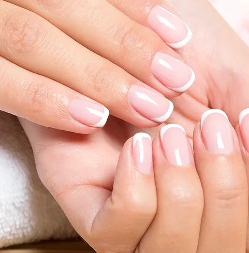 Close-up of hands with a French manicure, featuring neatly shaped nails with a pale pink base and white tips. The hands are resting on a soft white surface.