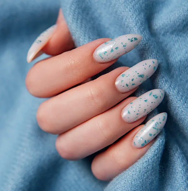 A close-up of a hand with long, almond-shaped nails. The nails are painted light blue with small, scattered dark blue speckles. The hand is resting on a soft, textured light blue fabric.