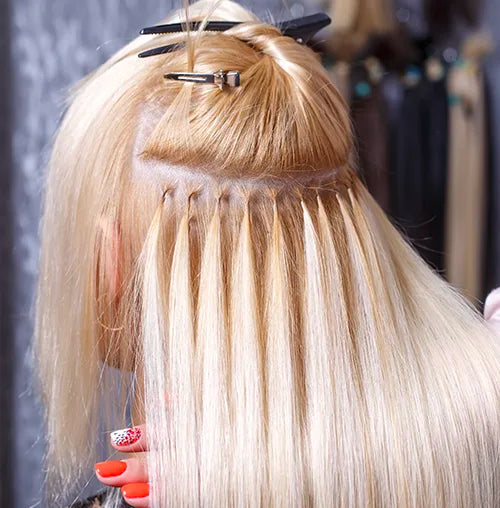 A person with light blonde hair having hair extensions applied using micro bead technique. Several strands are sectioned and secured with clips. The individual's nails are painted in red and white patterns.