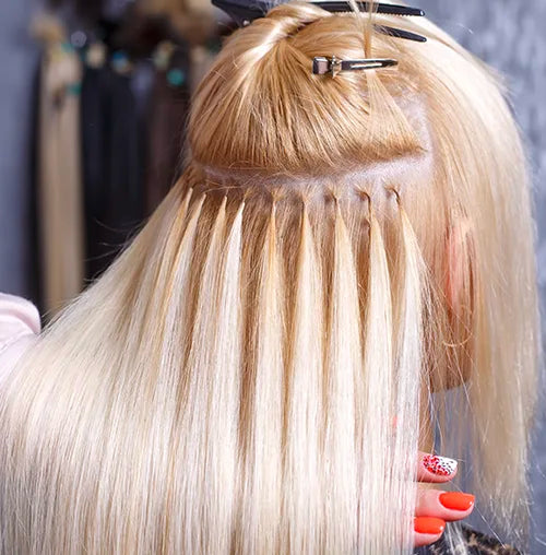 Close-up of blonde hair being styled with hair extensions using the micro loop technique. The extensions are neatly attached, with part of the natural hair clipped up. A person's hand with red and white patterned nails is visible at the bottom.