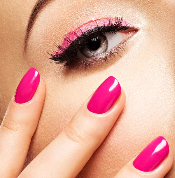 Close-up image of a person with bright pink eyeshadow and matching pink nail polish. The fingers are gently touching the cheek, highlighting the vibrant makeup and glossy nails against a soft skin tone.