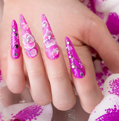 Close-up of a hand with long, pointed nails painted in bright pink, adorned with intricate 3D floral designs in shades of pink and white. The nails feature delicate pearl and gemstone accents. The background includes pink and white floral patterns.