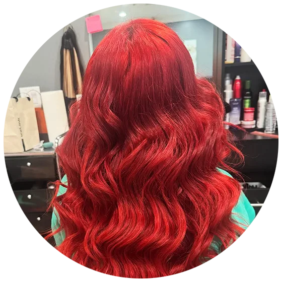 Person with long, wavy, vibrant red hair sitting in a salon chair, facing away. Shelves with hair products are visible in the background.