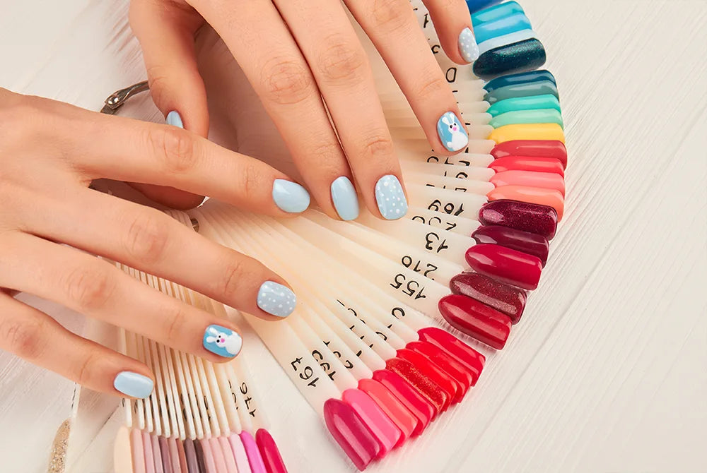 Hands with light blue nail polish and playful designs, featuring floral and polka dot patterns, rest on a fan of nail polish samples. The samples display a wide range of colors from pink to dark blue on a light wooden surface.